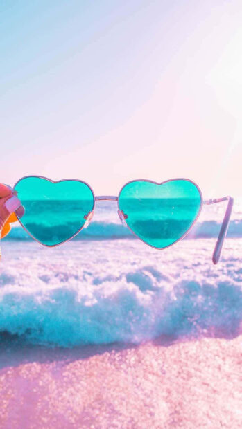 A hand holding heart shaped sunglasses with a summer ocean view through the lenses