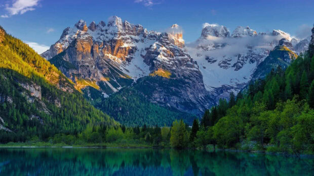 Snow capped spring mountain surrounded by lush green forest and calm lake reflecting the scenery