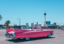 A classic pink convertible car driving on a sunny summer street with city buildings in the background