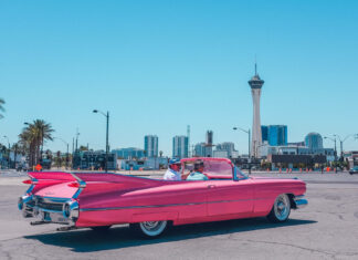 A classic pink convertible car driving on a sunny summer street with city buildings in the background