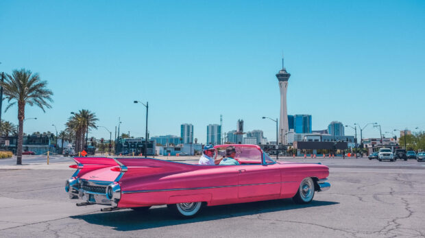 A classic pink convertible car driving on a sunny summer street with city buildings in the background