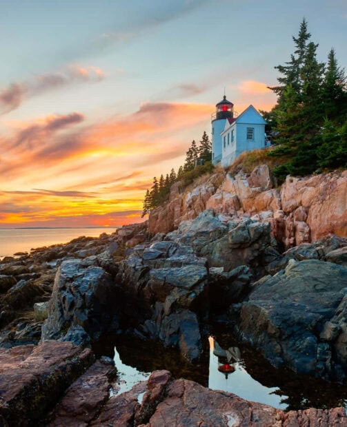 A rocky coastline with a lighthouse and pine trees during a summer sunset