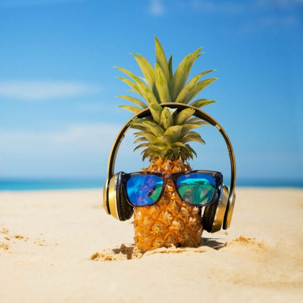 A pineapple wearing sunglasses and headphones on a sandy beach in summer
