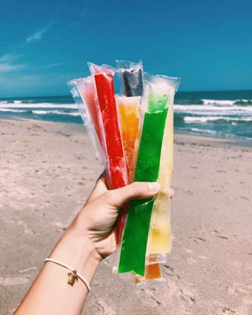 A hand holding colorful summer ice pops at the beach on a sunny day