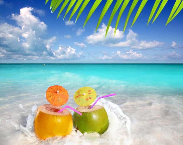 Two tropical coconuts with straws and paper umbrellas on the beach sand near turquoise ocean water