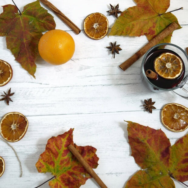 Mulled wine, spices, and autumn leaves arrange on a white wooden surface, featuring cinnamon, star anise, oranges, and colorful maple leaves