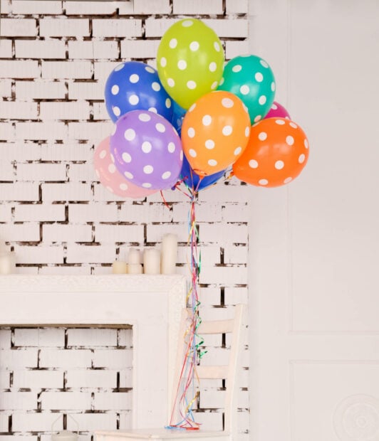 A vibrant bunch of polka-dot balloons floats, tied with colorful ribbons, decorating a bright room with a white brick fireplace