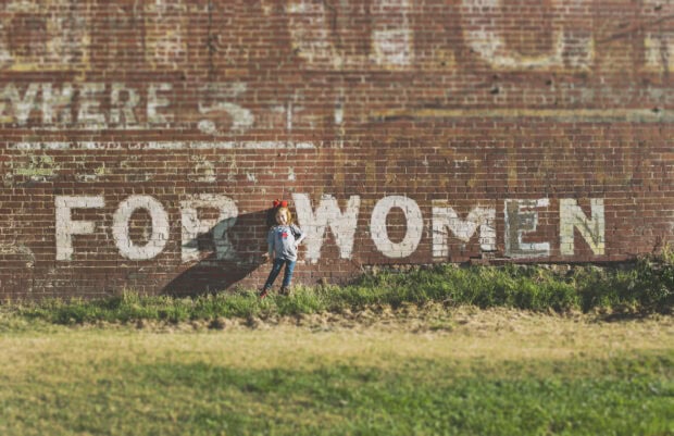 Young child stands before a weathered brick wall displaying large, faded white text 