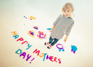 A small child stands on a white floor, colorful painted letters spell a greeting, playful drawings surround her