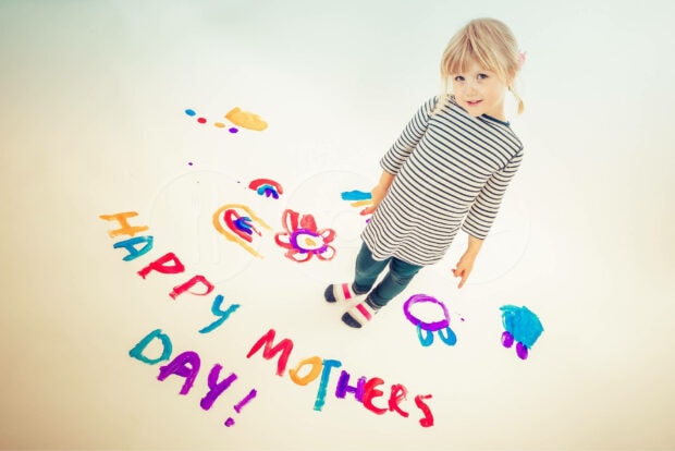 A small child stands on a white floor, colorful painted letters spell a greeting, playful drawings surround her