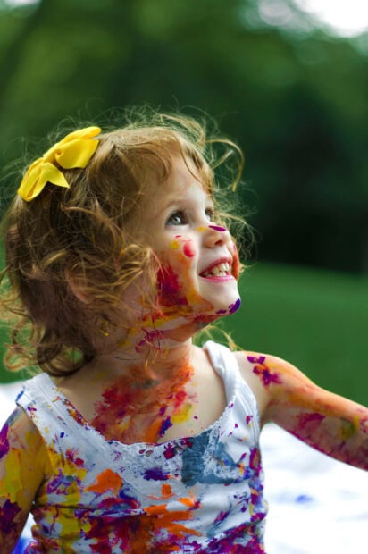 A toddler girl smiles brightly, looking up, her face and clothes covered in vibrant colorful paint during a playful outdoor activity