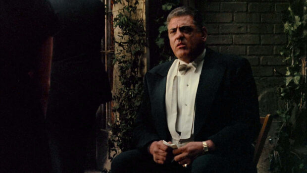A man in a tuxedo sits intently, hands clasped, against a brick wall adorned with green ivy
