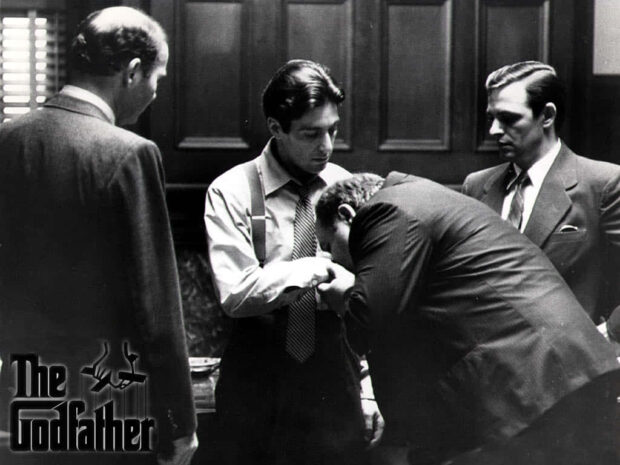 A man bows, kissing Michael Corleone's hand in a gesture of profound respect and allegiance, surrounded by other figures observing
