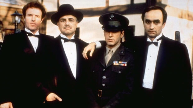 Four men standing side-by-side, three wearing tuxedos and one dressed in a military uniform, presenting a formal gathering or family moment