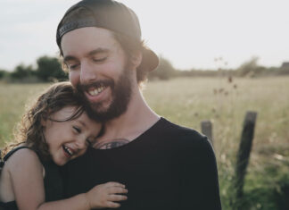 A bearded man embraces a young girl, both smiling warmly in a sunlit outdoor field