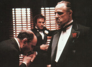 A man bows and kisses the hand of Vito Corleone, while Sonny Corleone observes in the background