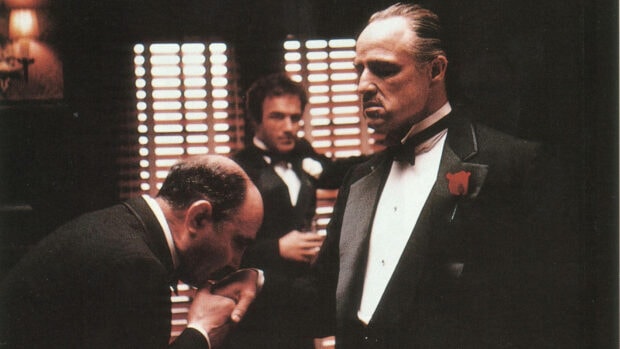 A man bows and kisses the hand of Vito Corleone, while Sonny Corleone observes in the background