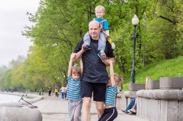 A man carries a young boy on his shoulders while two girls hang from his arms, walking along a path in a vibrant green park beside a body of water