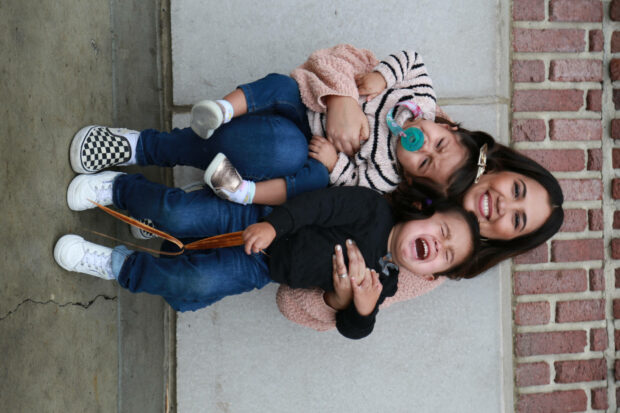 Woman lies down, two children pile on top, all laugh together by a brick wall
