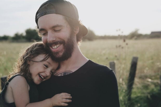 Man embraces child, both smiling brightly under a warm sunlit sky in a golden field
