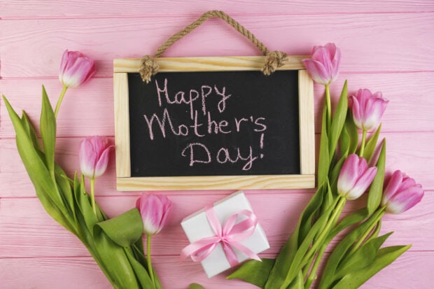 Pink tulips and a gift box flank a blackboard displaying a chalk message on a pink wooden surface