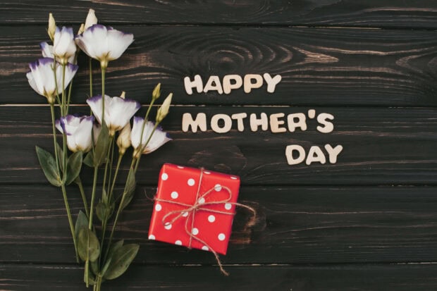 White and purple flowers accompany a polka-dotted gift box and wooden letters spelling a greeting on a dark plank surface