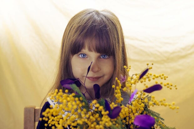 Young girl with blue eyes gazes forward, partially framed by bright yellow mimosa flowers and purple leaves