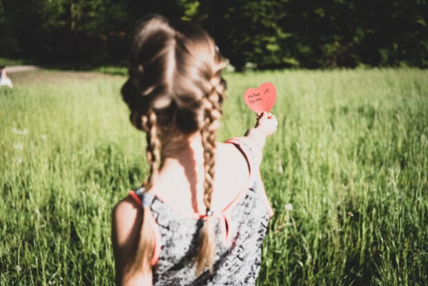 A young girl with braided hair stands in a sunny green field, holding a red heart-shaped card upwards