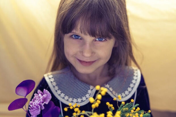 Young girl gazes forward with bright blue eyes, softly smiling while holding an assortment of purple and yellow flowers