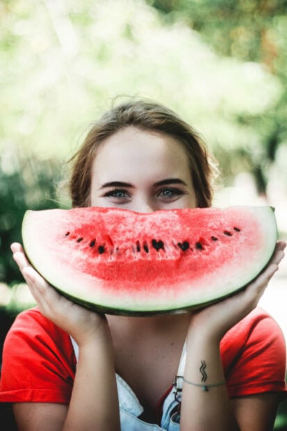 A young woman holds a large slice of watermelon, covering her mouth and nose, revealing only her joyful, smiling eyes against a soft green background