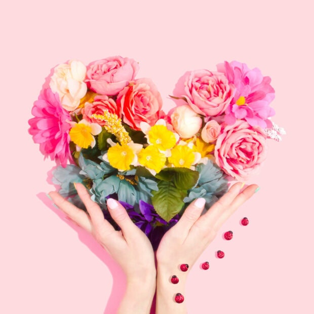 Hands cradle a vibrant heart-shaped bouquet of flowers against a pink background