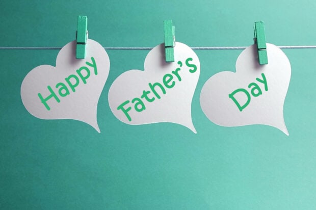Three white heart-shaped paper cutouts hang on a string with green clothespins, displaying 'Happy Father's Day' in green script against a teal background