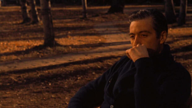Michael Corleone sits alone, appearing deeply thoughtful, hand resting on his chin amidst a dimly lit, autumn forest