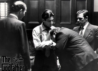 Michael Corleone accepts a man bowing and kissing his hand, surrounded by observing figures in a black and white scene