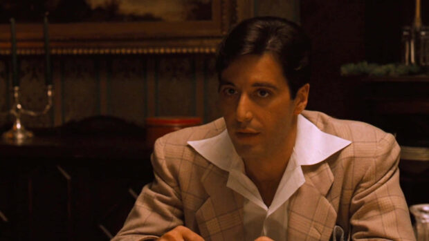 Michael Corleone sits indoors, wearing a plaid jacket and white shirt, his gaze focused and intense