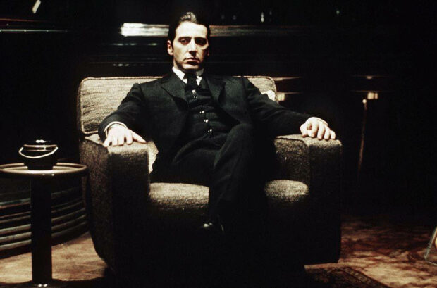 Michael Corleone sits upright in an armchair, directly facing forward, his expression resolute in a dimly lit, powerful setting