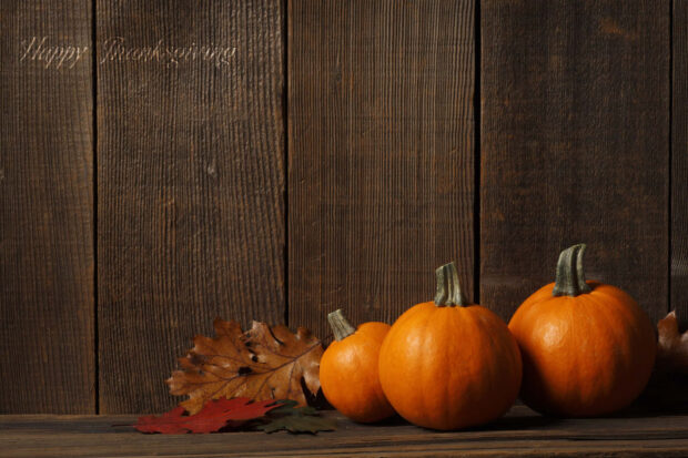 Three small orange pumpkins rest beside dried autumn leaves on a rustic wooden surface against a dark plank background