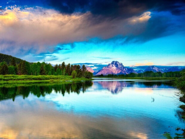 Majestic mountains reflect in serene lake, surrounded by dense green forest under a vivid sky