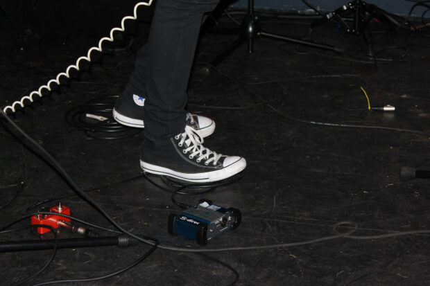 A person stands wearing black high-top sneakers on a dark stage floor, surrounded by coiled audio cables and music effects pedals