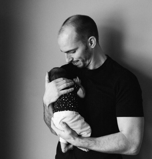 Man gently cradles infant, gazing down with a soft smile in a black and white portrait