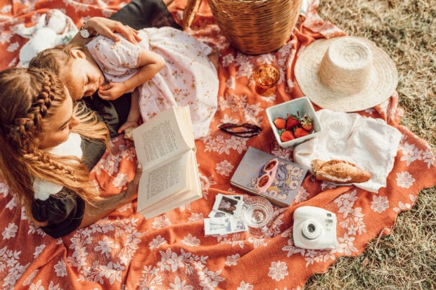 Two women relax on a picnic blanket, one reading a book, the other peacefully sleeping