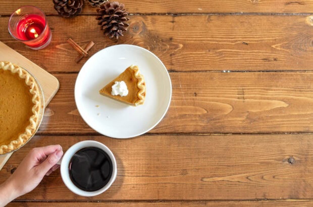 A slice of pumpkin pie topped with whipped cream, a full pie, and a cup of coffee rest on a rustic wooden table alongside pinecones, cinnamon sticks, and a lit candle, with a hand holding the coffee