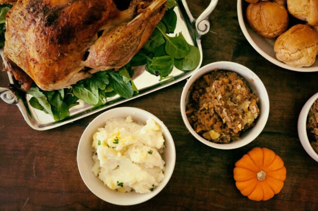 A perfectly roasted turkey on a silver platter, bowls of mashed potatoes and stuffing, and dinner rolls are arranged on a dark wooden table, accompanied by a small decorative pumpkin