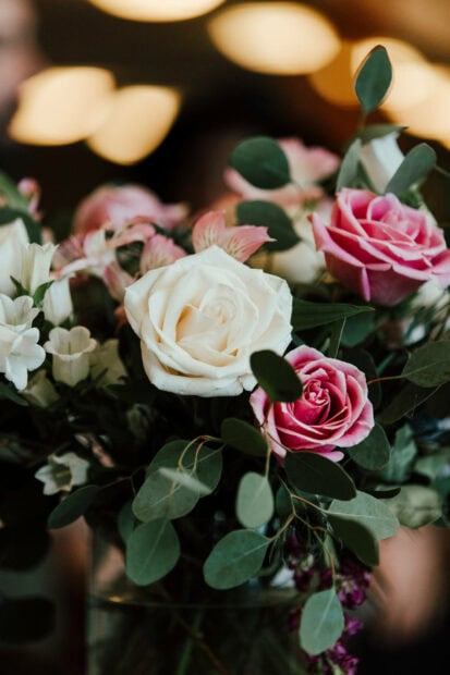 A lush bouquet features blooming white and pink roses nestled among deep green foliage, with warm, blurred lights in the background