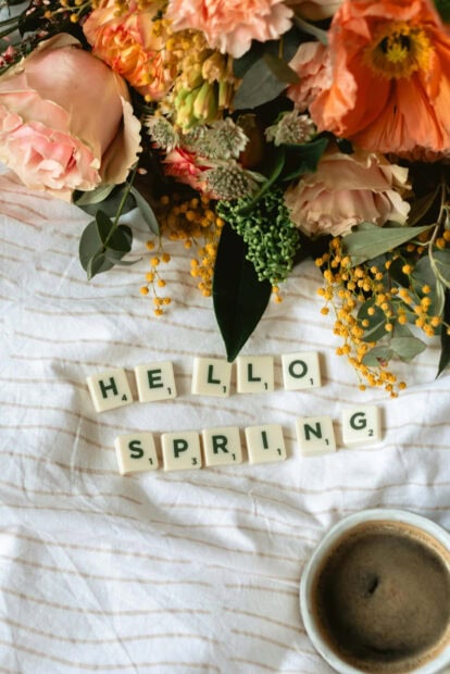 Scrabble tiles spell out a greeting on a textured sheet, surrounded by fresh orange and pink blossoms with a cup of coffee