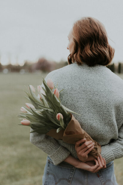 Woman stands with back turned, hiding a bouquet of pink tulips wrapped in paper