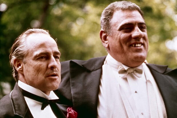 Don Vito Corleone stands beside Luca Brasi, Brasi smiles broadly, Corleone appears thoughtful in formal tuxedos outdoors