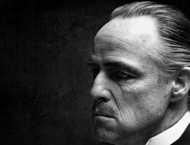 Close-up profile of Marlon Brando as Vito Corleone gazes pensively, showing weathered features against a dark, textured backdrop