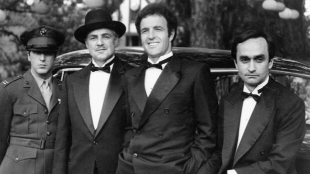 Four men stand together in formal attire, two wearing tuxedos, one in a fedora, and another in military uniform, beside a classic car