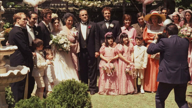 Wedding guests gather outdoors, posing for a group photograph as a photographer captures the moment